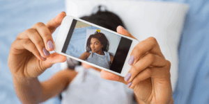 A cellphone close up with an image of a young African American woman on it and the same woman behind blurred, taking a selfie
