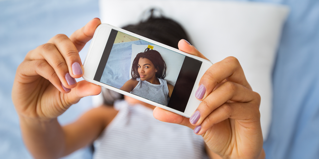 A cellphone close up with an image of a young African American woman on it and the same woman behind blurred, taking a selfie